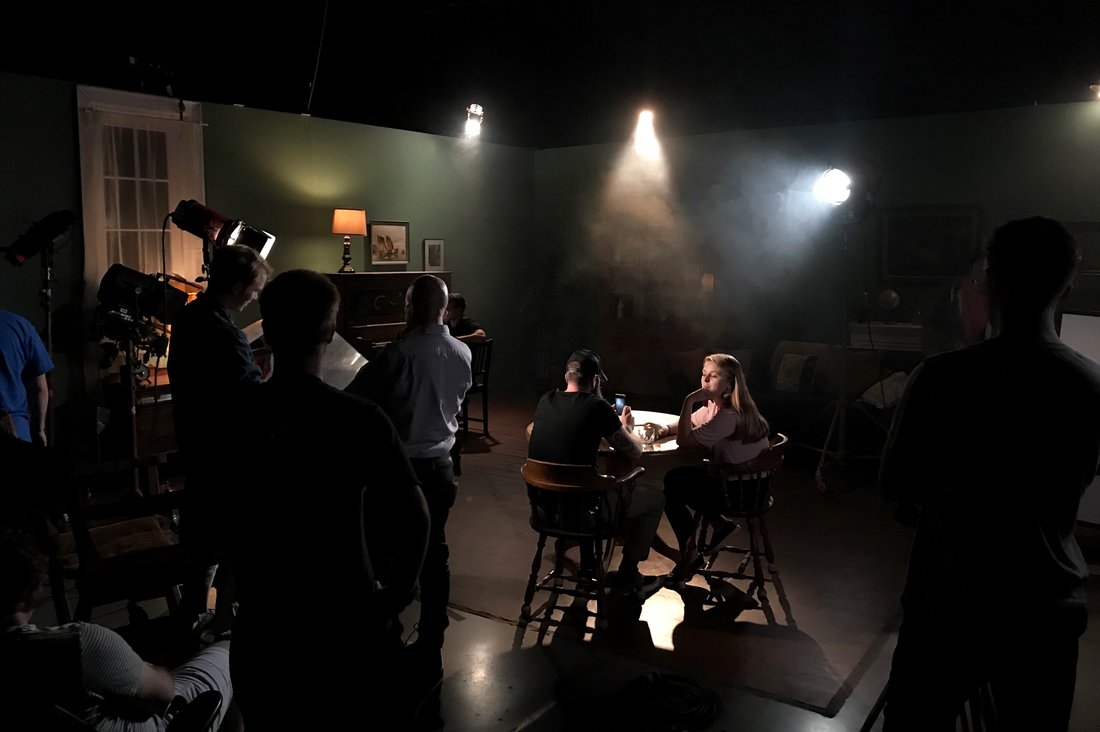 Two actors sit a dimly-lit table surrounded by members of a film crew taping a scene