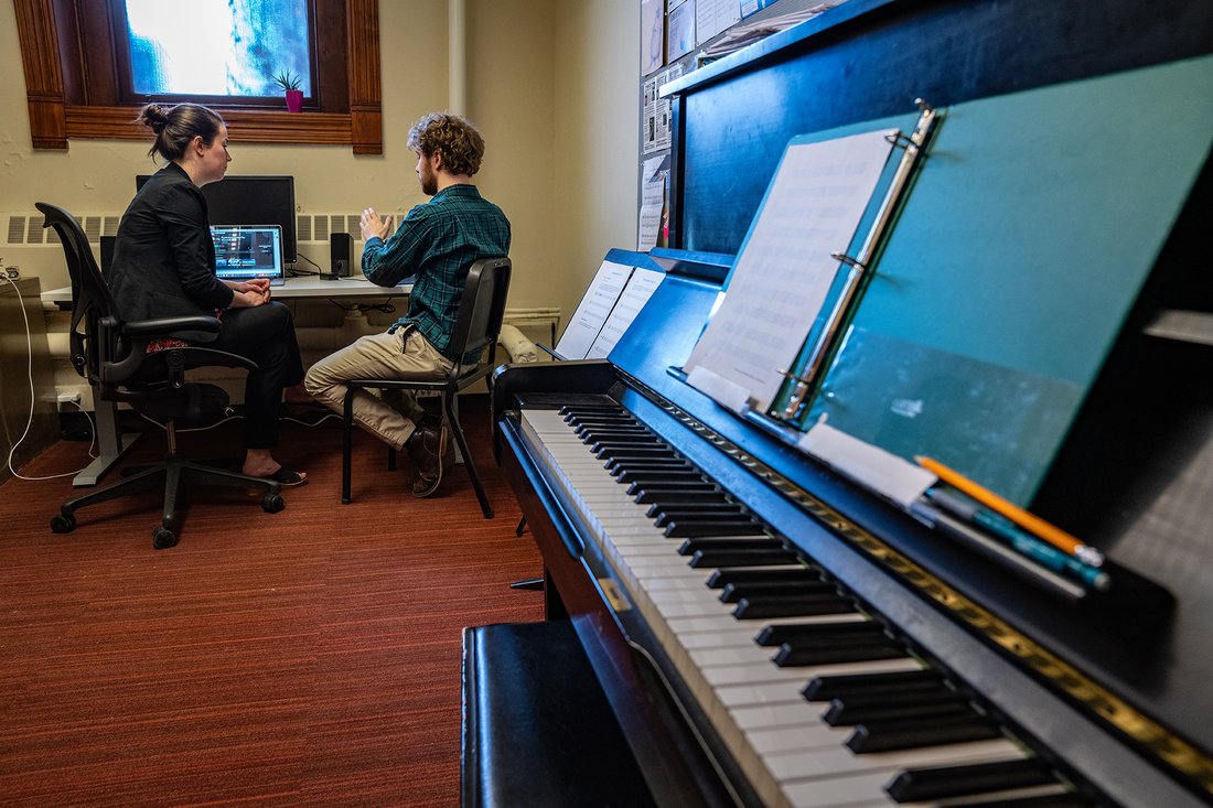 A teacher and student sit at a desk in an office with a piano