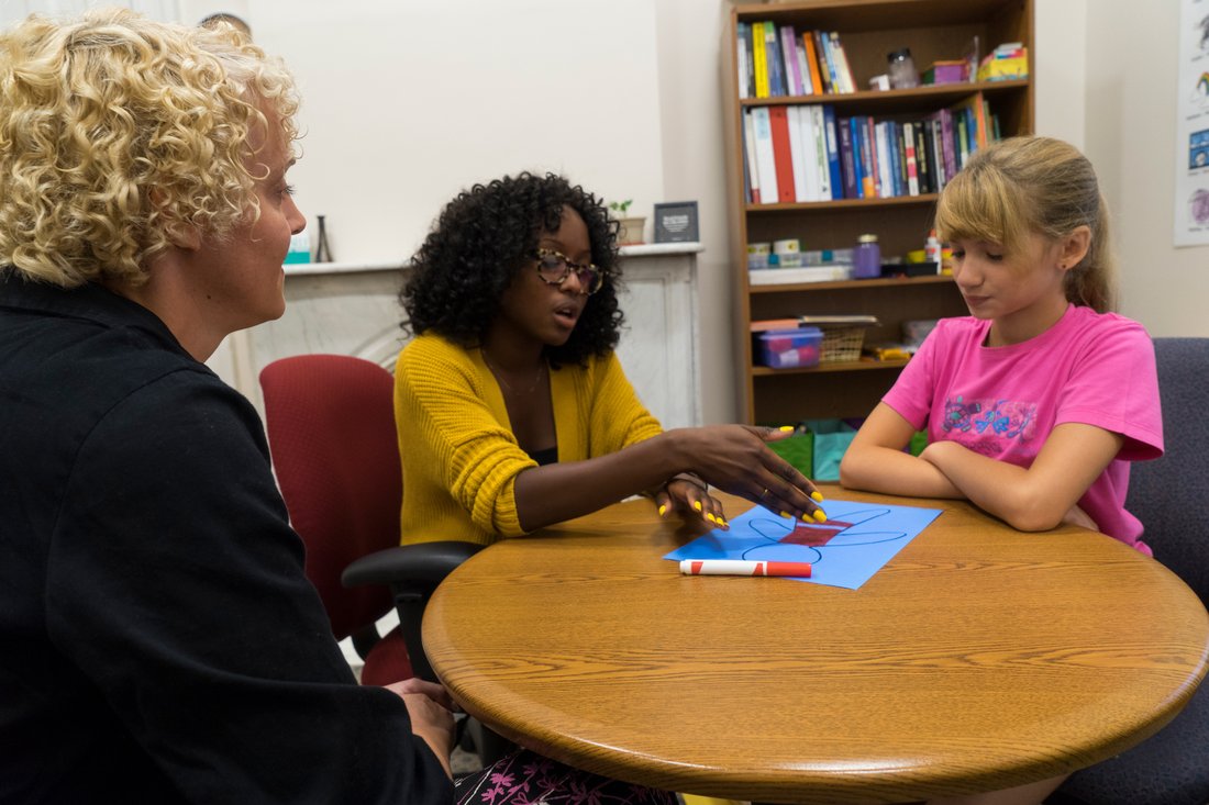 A student therapist sits with an adult and a child at a table. They talk about the child