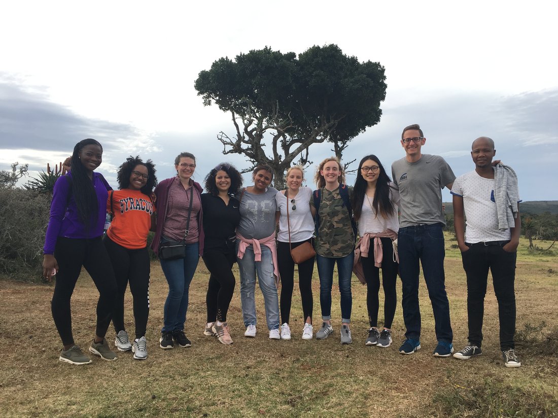 nine students and staff pose in a line for the camera in front of a large tree in south africa