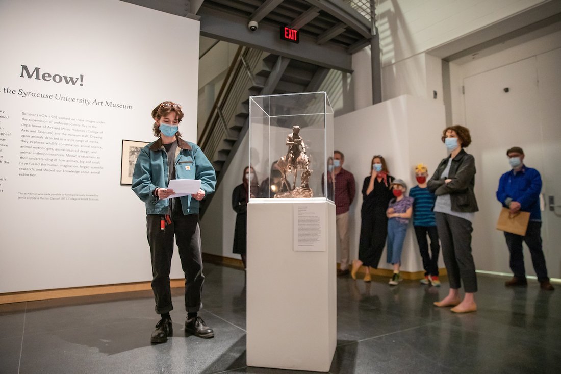 A student stands next to a statue while reading from a document inside a Syracuse University Art Museum gallery.