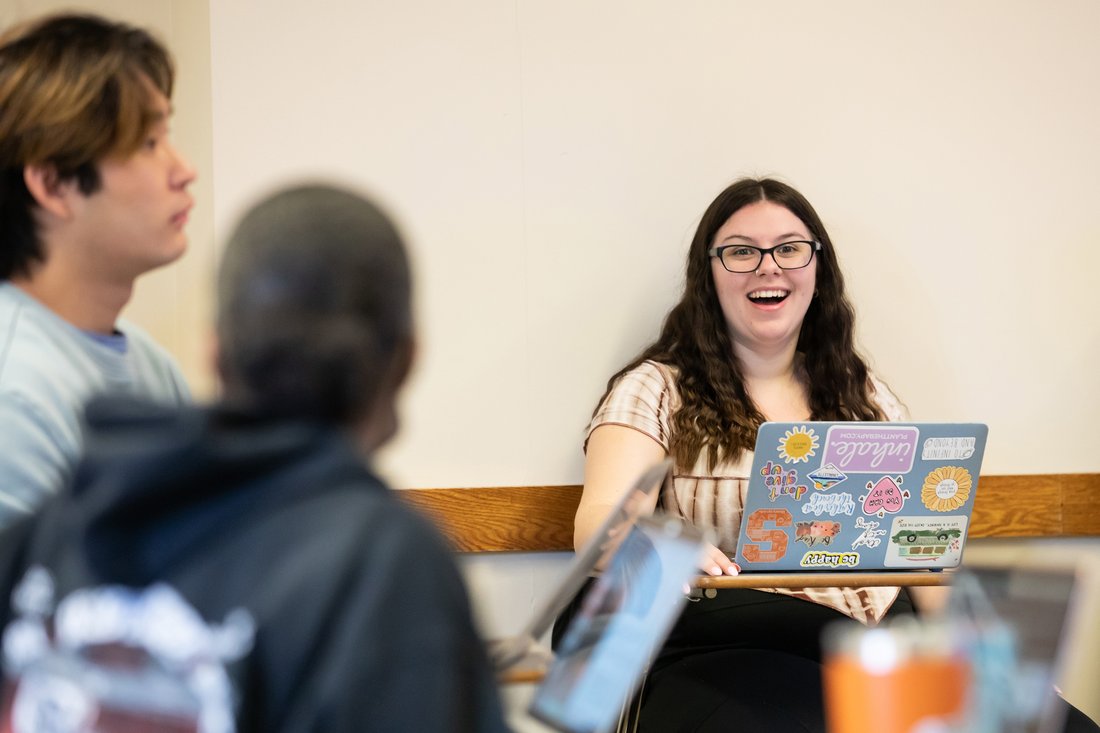 a student smiles in class while on a laptop covered in stickers and talking to two other students