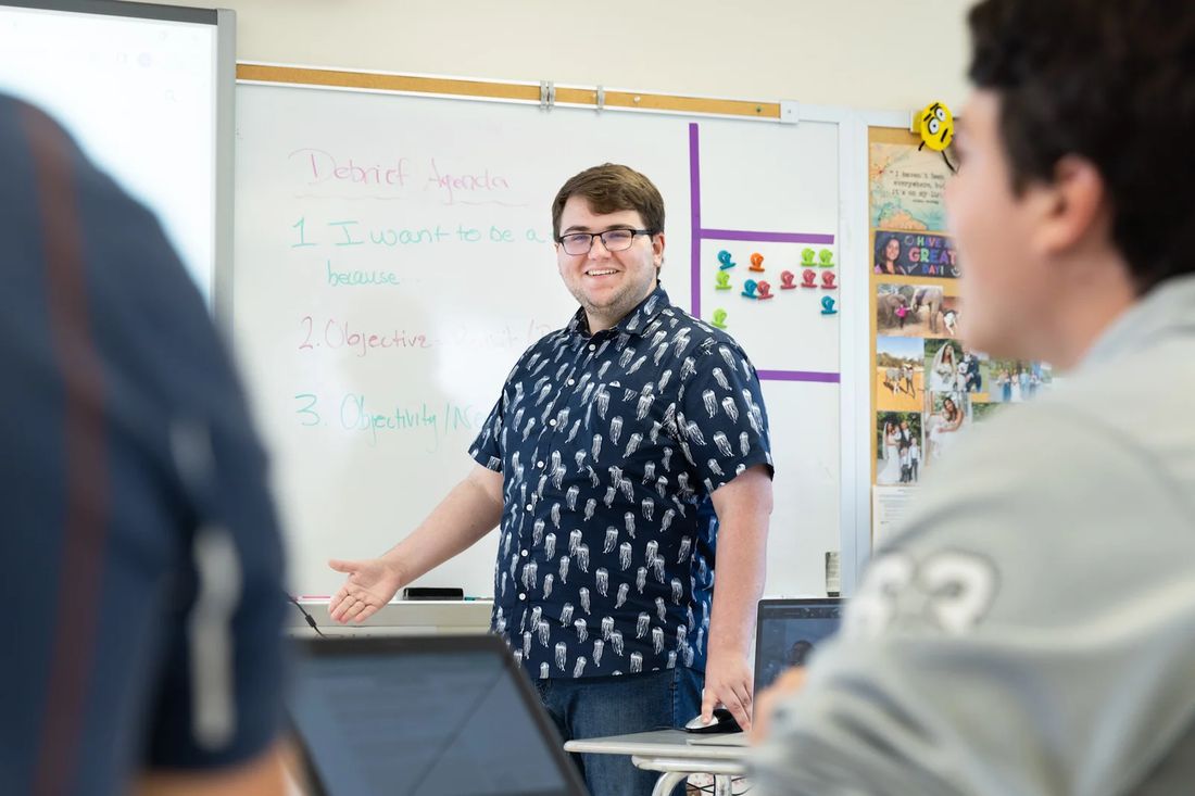 a male student at a whiteboard teaches several high school students